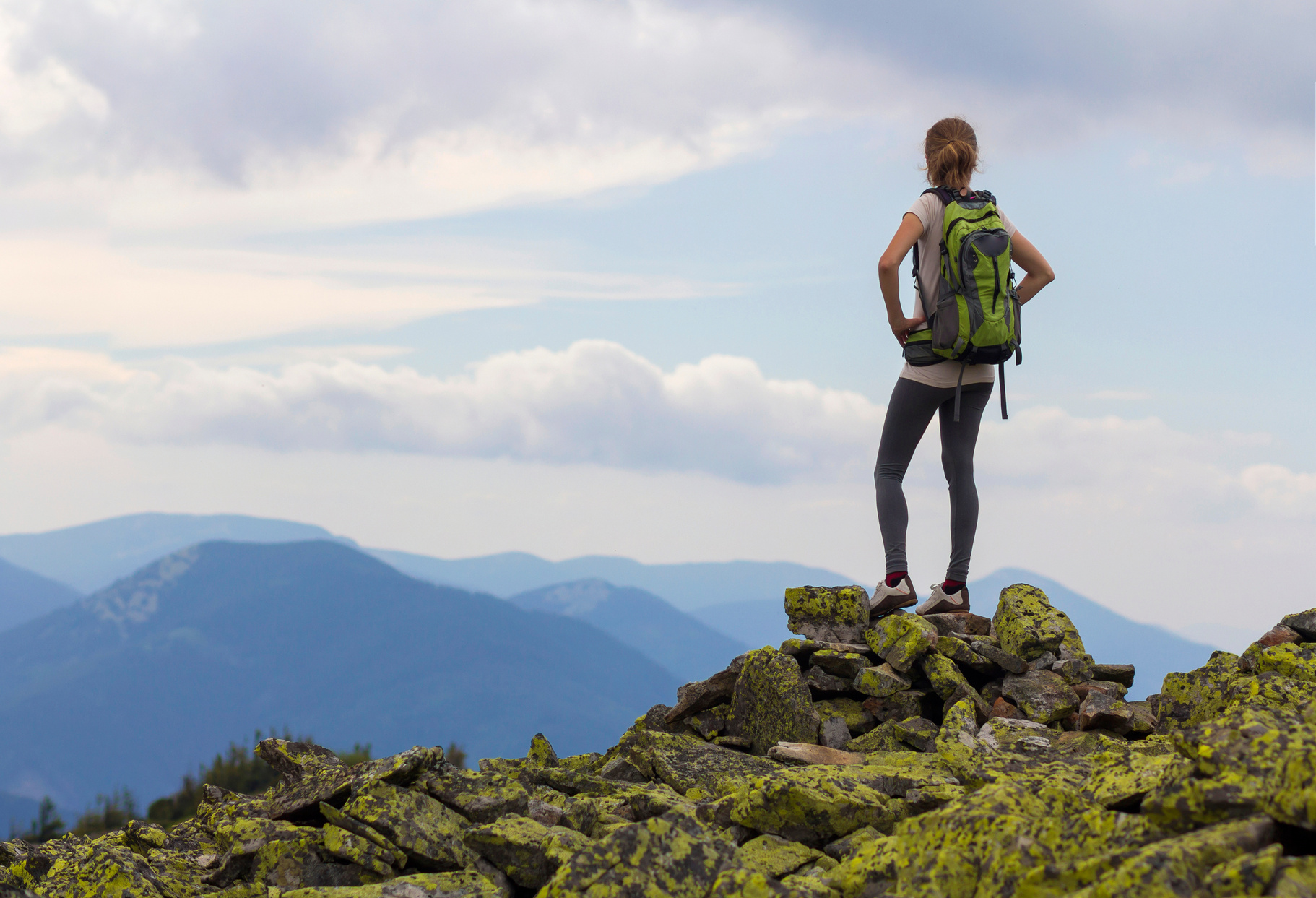 Back view of young slim girl with backpacks standing on rocky mountain top against bright blue morning sky enjoying foggy mountain range panorama. Tourism, traveling and healthy lifestyle concept.